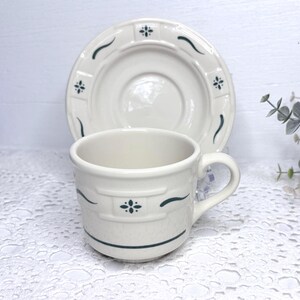 May include: A cream-colored ceramic cup and saucer set featuring a dark green floral motif. The cup has a rounded handle and a horizontal band near the base, while the saucer displays a matching pattern of small floral accents and curved lines.