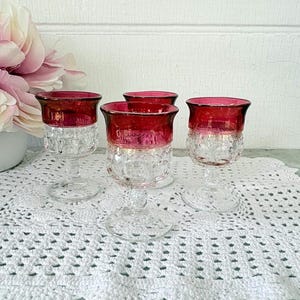 May include: Four vintage wine glasses with ruby red rims and clear, textured glass bodies. The glasses are arranged on a white lace doily, with a bouquet of pink flowers in the background.