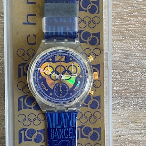 May include: A Swatch chronograph watch with a blue face, gold accents, and a blue strap. The watch face features multiple dials and the numbers 1894 and 1994. The strap has the text "STOCKHOLM LONDON ATLANTA BARCELONA SEOUL SARAJEVO".