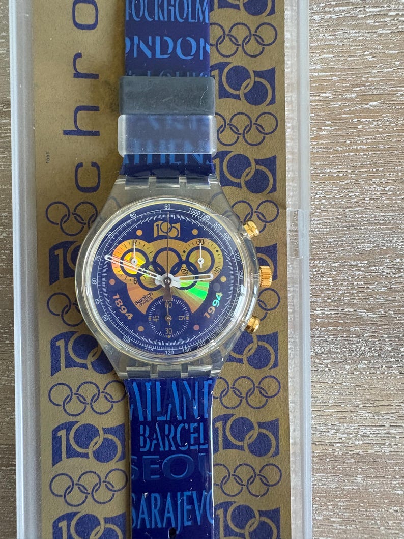 May include: A Swatch chronograph watch with a blue face, gold accents, and a blue strap. The watch face features multiple dials and the numbers 1894 and 1994. The strap has the text "STOCKHOLM LONDON ATLANTA BARCELONA SEOUL SARAJEVO".