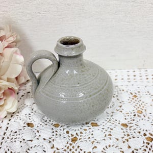 May include: A small, gray ceramic jug with a handle and a narrow neck. The jug has a rounded body with horizontal etched lines and a slightly textured surface. The handle curves gracefully from the body to the neck. The jug is likely vintage or antique.