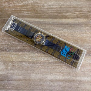 May include: A Swatch Chrono watch in its original packaging. The watch has a blue strap with gold accents and a gold-toned watch face. The packaging features the Olympic rings and the text "100 Years of the Olympic Movement".