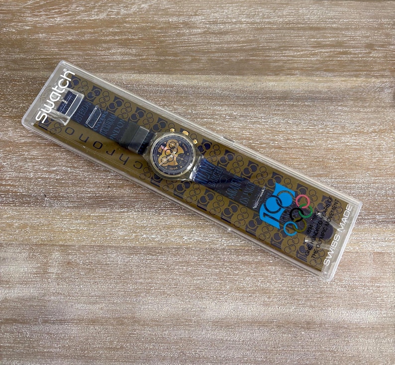 May include: A Swatch Chrono watch in its original packaging. The watch has a blue strap with gold accents and a gold-toned watch face. The packaging features the Olympic rings and the text "100 Years of the Olympic Movement".