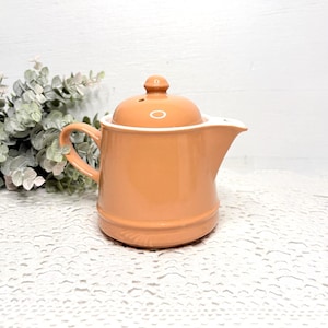May include: A peach-colored ceramic teapot with a smooth, glossy finish. The teapot features a rounded lid with a small knob handle, a curved side handle, and a short, wide spout. The body of the teapot has a slightly tapered cylindrical shape.