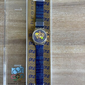 May include: A Swatch chronograph watch in its original packaging. The watch has a blue strap with Olympic rings and city names. The watch face has gold accents and the Olympic rings logo. The packaging is clear plastic.