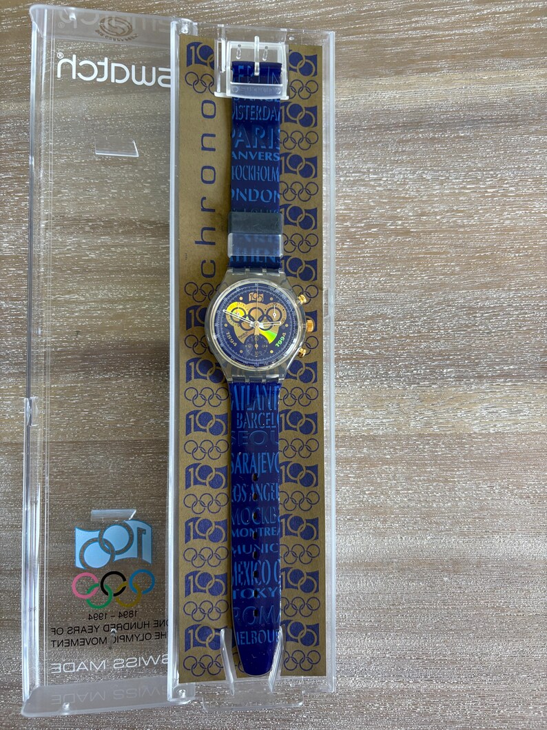 May include: A Swatch chronograph watch in its original packaging. The watch has a blue strap with Olympic rings and city names. The watch face has gold accents and the Olympic rings logo. The packaging is clear plastic.