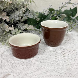 May include: Two brown ceramic ramekins with textured exteriors and white interiors. One is a low, round shape, and the other is a taller, cylindrical shape. The ramekins are set on a white lace tablecloth, with greenery in the background.