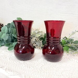 May include: Two ruby red glass vases with a bulbous base and a flared top. The vases have a textured pattern on the lower half and a smooth, glossy finish. They are displayed on a white lace surface with greenery in the background.