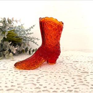 May include: A vibrant orange glass boot figurine featuring a detailed pressed starburst and diamond pattern across its surface. The boot has a scalloped top edge and a small heel, crafted from translucent glass that catches the light.