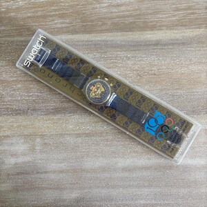 May include: A Swatch chronograph watch in its original packaging. The watch has a black strap with gold accents and a gold-toned face. The packaging features the Olympic rings logo and text: "100 Years of the Olympic Movement."