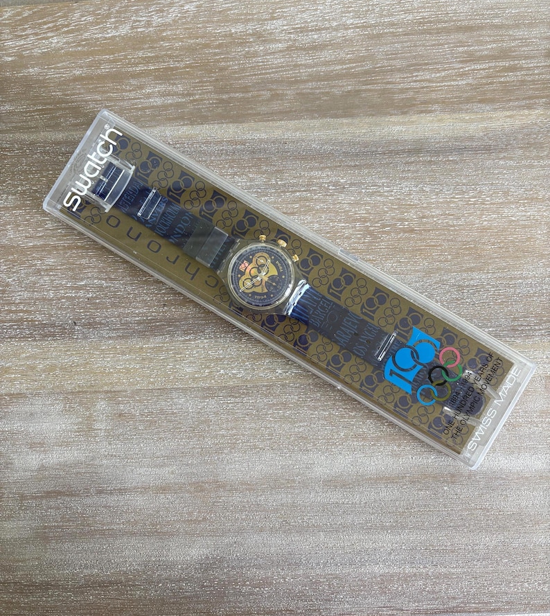 May include: A Swatch chronograph watch in its original packaging. The watch has a black strap with gold accents and a gold-toned face. The packaging features the Olympic rings logo and text: "100 Years of the Olympic Movement."