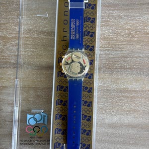 May include: A blue Swatch watch with a blue strap, gold-coloured face, and clear plastic case. The watch features the text "REMEMBER SARAJEVO" and Olympic rings. The watch is in its original packaging.