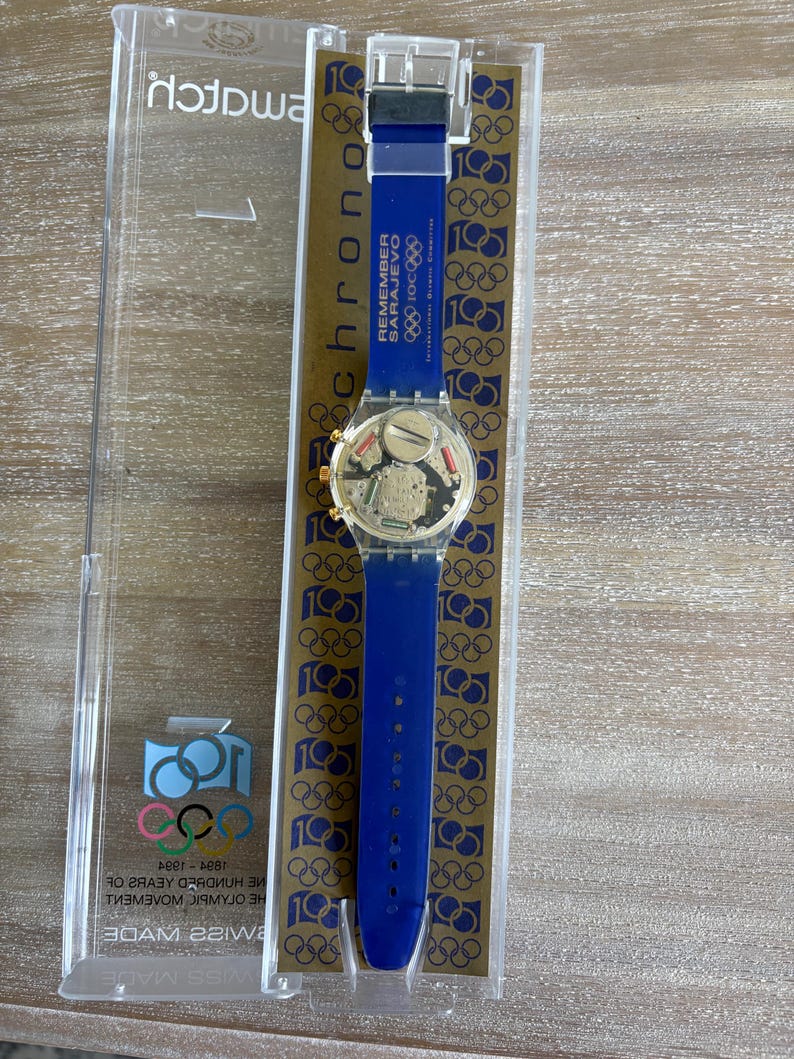 May include: A blue Swatch watch with a blue strap, gold-coloured face, and clear plastic case. The watch features the text "REMEMBER SARAJEVO" and Olympic rings. The watch is in its original packaging.