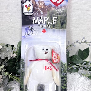 May include: A white plush Maple the Bear Beanie Baby toy in its original packaging. The packaging features a scenic background with mountains and the text "MAPLE THE BEAR". The bear has a red ribbon and a Canadian flag.