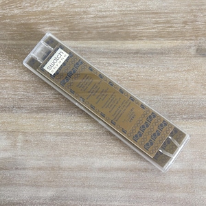 May include: A clear plastic case containing a gold-coloured watch component. The component features the text "Swatch Chrono" and Olympic rings. The case is rectangular and angled on a wooden surface.