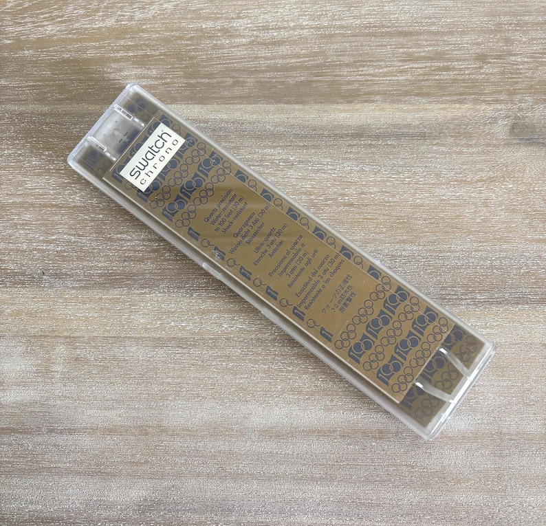 May include: A clear plastic case containing a gold-coloured watch component. The component features the text "Swatch Chrono" and Olympic rings. The case is rectangular and angled on a wooden surface.