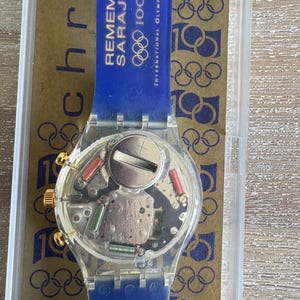 May include: A clear Swatch watch with a blue strap. The watch face reveals the internal components. The blue strap has the text "REMEMBER SARAJEVO" and Olympic rings. Gold-coloured accents are on the watch face.