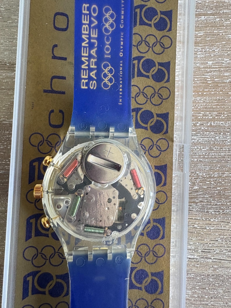 May include: A clear Swatch watch with a blue strap. The watch face reveals the internal components. The blue strap has the text "REMEMBER SARAJEVO" and Olympic rings. Gold-coloured accents are on the watch face.
