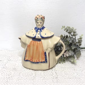 May include: A ceramic teapot shaped like a person wearing a bonnet, a cape with blue polka dots, and an orange apron. The figure has a painted face, a blue bow at the neck, and a handle on the side. The teapot is cream-colored with blue trim.