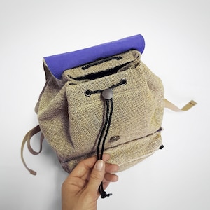 Könnte beinhalten: Ein kleiner, beiger Jutesack mit lila Futter und schwarzem Kordelzug. Der Rucksack hat eine kleine Tasche auf der Vorderseite und einen einzigen Riemen.