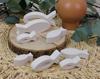 May include: A set of white wooden fish figurines arranged on a round woven mat with a brown vase in the background. The fish are of varying sizes and shapes, with some facing left and others facing right.