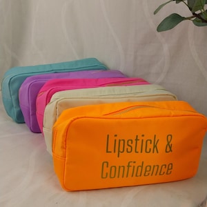 Könnte beinhalten: Eine Reihe farbenfroher Kosmetiktaschen in Türkis, Lila, Pink, Creme und Orange. Auf der orangefarbenen Tasche steht der Text "Lipstick & Confidence". Jede Tasche hat einen goldenen Reißverschluss.