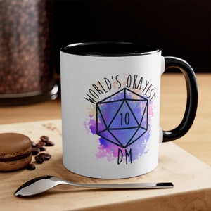 May include: White ceramic mug with a black interior and handle. The mug features the text "WORLD'S OKAYEST DM" and a geometric design with the number 10. The design is set against a watercolor-style background with purple, blue, and pink hues.