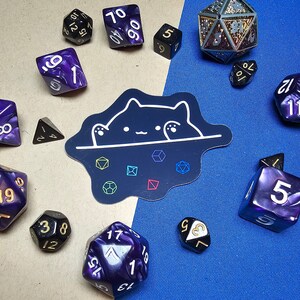 May include: A collection of purple, black, and gold dice with white numbers. The dice are arranged in a circle around a sticker of a cat peeking over a wall. The sticker has a black background and white outline.