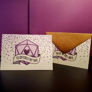 May include: A card with a purple 20-sided die with a heart inside and the text "I'd critically hit that". The card is surrounded by small purple hearts. The card is in a brown envelope with the text "You're my roll mate".