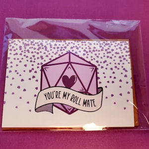 May include: A white card with a purple 20-sided die with a heart in the center and a banner that says "You're my roll mate". The background is covered in small purple hearts.