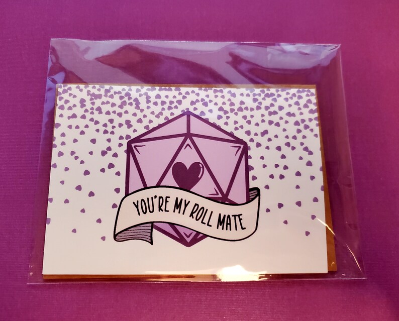 May include: A white card with a purple 20-sided die with a heart in the center and a banner that says "You're my roll mate". The background is covered in small purple hearts.