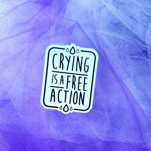 May include: A white sticker with black text on a purple and blue tulle background. The text reads "Crying is a free action".