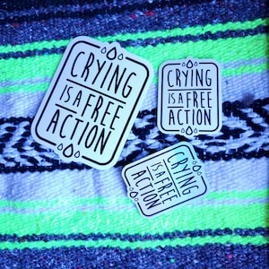 May include: Three white stickers with black text that reads "Crying is a free action" on a colorful striped background.