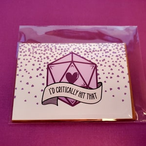 May include: A white card with a pink and purple dice with a heart in the center. The card has a banner that says "I'd critically hit that". The card is surrounded by a pattern of small pink hearts.