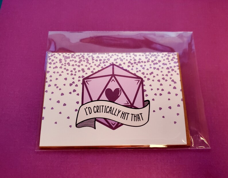 May include: A white card with a pink and purple dice with a heart in the center. The card has a banner that says "I'd critically hit that". The card is surrounded by a pattern of small pink hearts.
