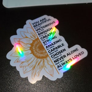 May include: A sticker featuring a sunflower design with a rainbow iridescent finish. The sticker includes inspirational text with bible verses, such as "You are beautiful", "Victorious", "Enough", "Created", "Strong", "Amazing", "Capable", "Chosen", "Never Alone", and "Always Loved".