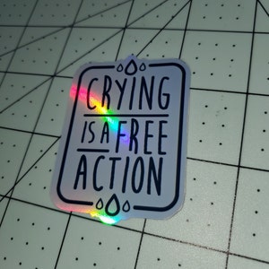 May include: A white sticker with a black border and black text that reads "Crying is a free action". The sticker has a holographic finish and is decorated with raindrops.