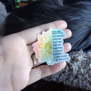 May include: A holographic sticker featuring a sunflower design and inspirational text. The text reads "You are beautiful, victorious, enough, created, strong, amazing, capable, chosen, never alone, always loved".