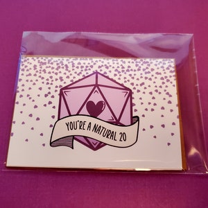 May include: A white card with a pink and purple 20-sided die with a heart in the center. The card has a gold border and a banner that reads "You're a natural 20". The background is covered in small pink hearts.