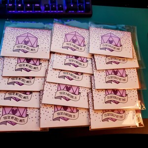 May include: A stack of 15 pink and purple greeting cards with a dice design and various messages, including "You're my roll mate", "You're a natural 20", and "I'd critically hit that".