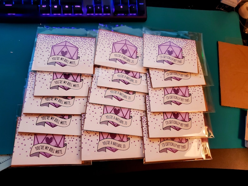 May include: A stack of 15 pink and purple greeting cards with a dice design and various messages, including "You're my roll mate", "You're a natural 20", and "I'd critically hit that".