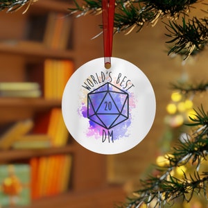 May include: A white, round ornament with the text "WORLD'S BEST DM" and a D20 die graphic. The die is blue with a "20" in the center, set against a watercolor-style purple and pink background. A red ribbon is attached for hanging.