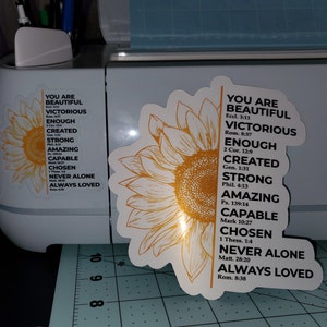 May include: A yellow sunflower with a white outline and a vertical list of positive affirmations with bible verses. The text reads: "You are Beautiful, Victorious, Enough, Created, Strong, Amazing, Capable, Chosen, Never Alone, Always Loved".