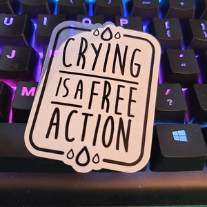 May include: A white sticker with black text and raindrop designs that reads "CRYING IS A FREE ACTION". The sticker is placed on a black keyboard with blue and purple lighting.
