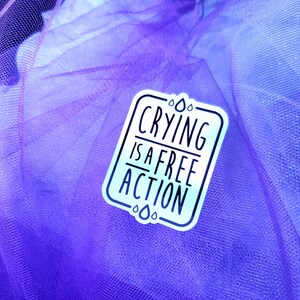 May include: A white sticker with a blue and purple ombre effect. The sticker has a black border and says "Crying is a free action" with raindrops around the text.