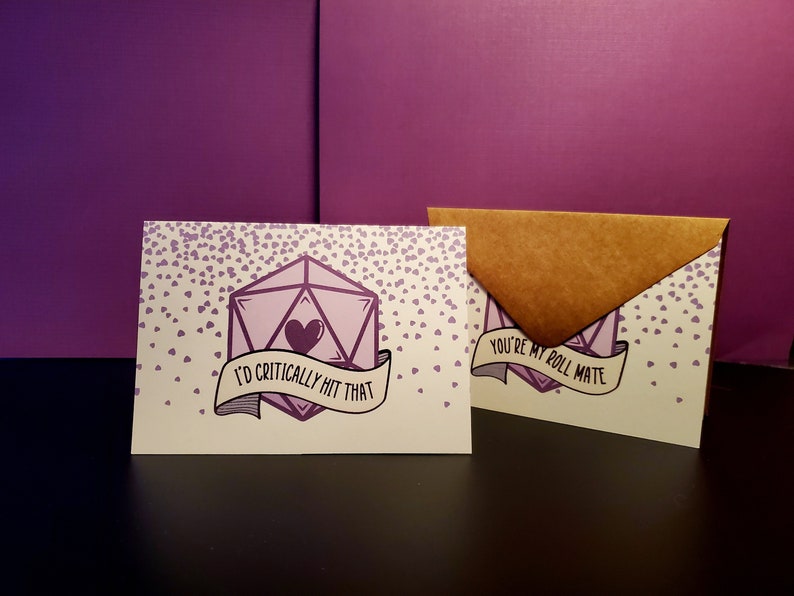 May include: A card with a purple 20-sided die with a heart inside and the text "I'd critically hit that". The card is surrounded by small purple hearts. The card is in a brown envelope with the text "You're my roll mate".