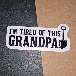 May include: A white sticker with black text that reads "I'M TIRED OF THIS GRANDPA" with a shovel graphic. The sticker is on a dark green and tan background.
