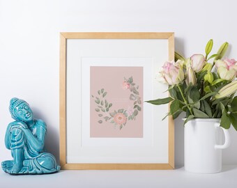 May include: A framed print featuring a floral wreath in pastel pink and green. The print is in a light wood frame, alongside a turquoise Buddha statue and a white enamelware vase with pink and white roses and lilies.