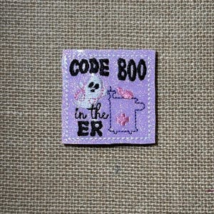 May include: A square lavender patch with a glittery finish featuring black embroidered text that reads CODE BOO in the ER. The design includes a white ghost character, a pink medical monitor, and a small pink cross symbol stitched onto the fabric.