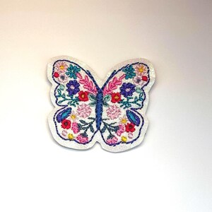 May include: A butterfly-shaped embroidered patch featuring a white background with a dense floral pattern in shades of red, pink, purple, blue, and green. The butterfly has a dark blue central body and a dark blue stitched border outline.